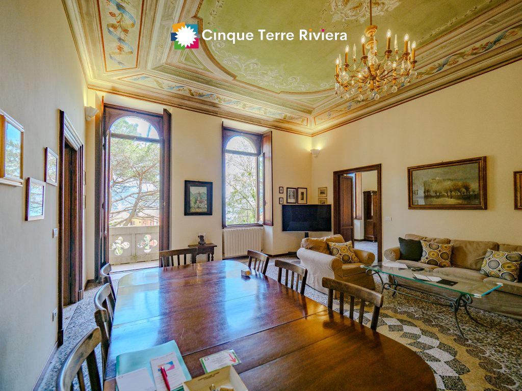 Salón-comedor de la finca Montale con techo pintado con motivos ornamentados, lámpara de araña de cristal, ventanas en arco, mesa de comedor de madera y una marca de agua «Cinque Terre Riviera» en la parte superior central.