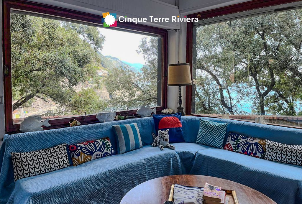 Sofá modular curvo de color azul brillante con cojines de estampados coloridos en un salón bañado por la luz del sol, con grandes ventanales que dan a la vegetación y al mar; diseño de interiores Cinque Terre Riviera.