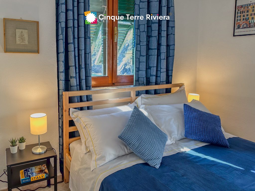 Escena del dormitorio de Teresa con una estructura de cama de madera, sábanas blancas con ribetes amarillos, cojines azules, cortinas azul oscuro y lámparas de noche cálidas que crean un ambiente costero y acogedor.