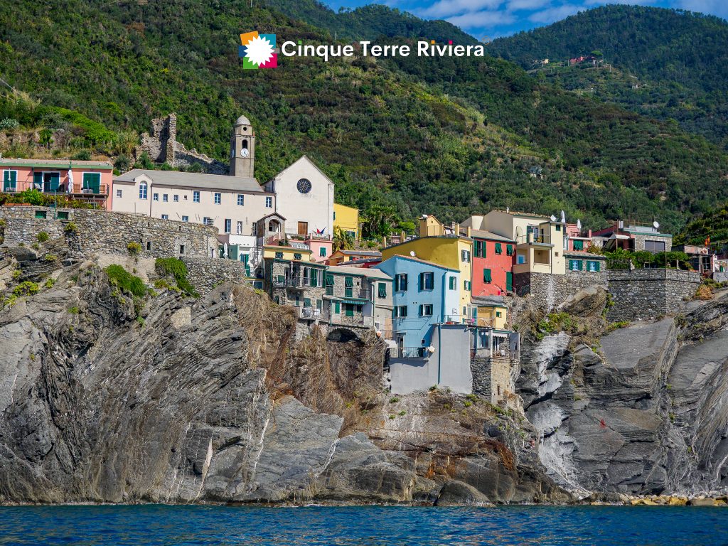 Casas de colores pastel encaramadas en un acantilado escarpado sobre el mar azul, con la torre de una iglesia y laderas verdes en Cinque Terre, Liguria, Italia.