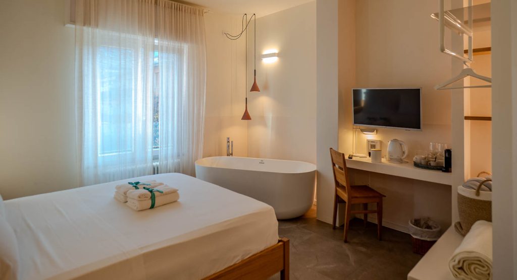 Habitación de hotel de lujo con ropa de cama blanca y cortinas translúcidas, una bañera exenta junto a una ventana, un televisor montado en la pared y un escritorio minimalista.