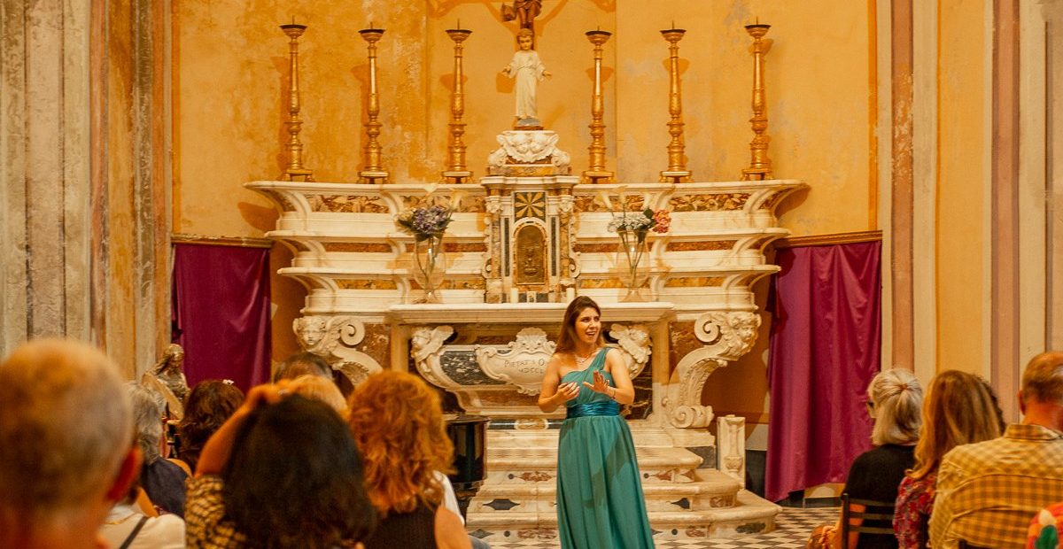 Una soprano con un vestido verde azulado actúa en los Conciertos de Ópera de Vernazza, en el ornamentado Oratorio de la Santissima Trinità, ante cortinas rojas.