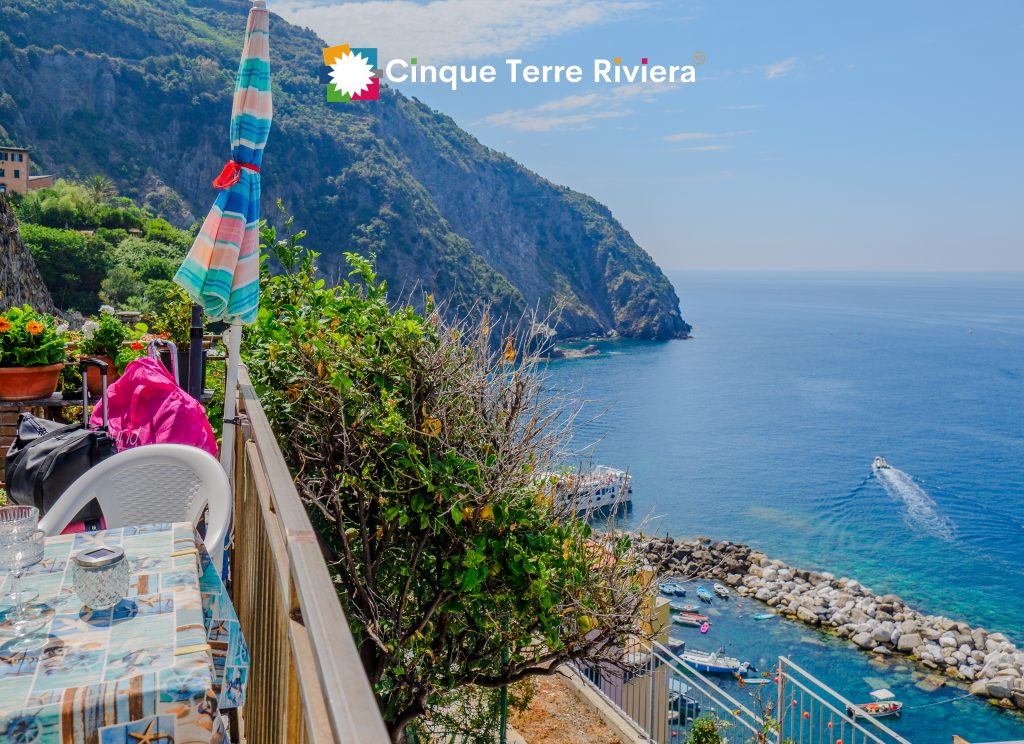 Una sombrilla de patio a rayas se encuentra junto a una terraza junto al mar con vistas a empinados acantilados verdes, un puerto rocoso y agua azul tranquila bajo un cielo despejado.