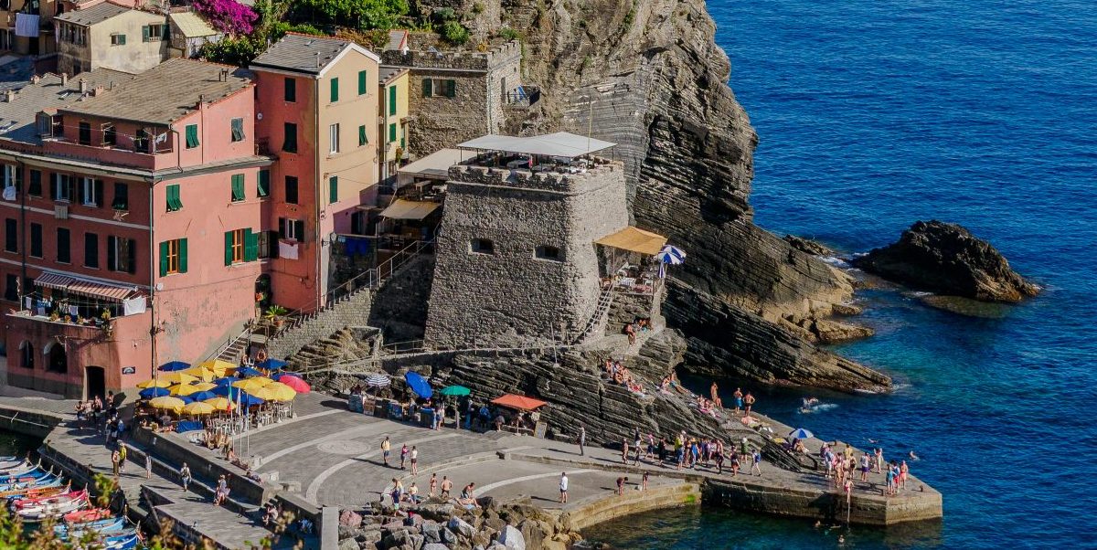 El puerto de Vernazza en Cinque Terre muestra casas pastel, una torre de piedra, sombrillas y gente junto al azul mar de Liguria en una costa rocosa.