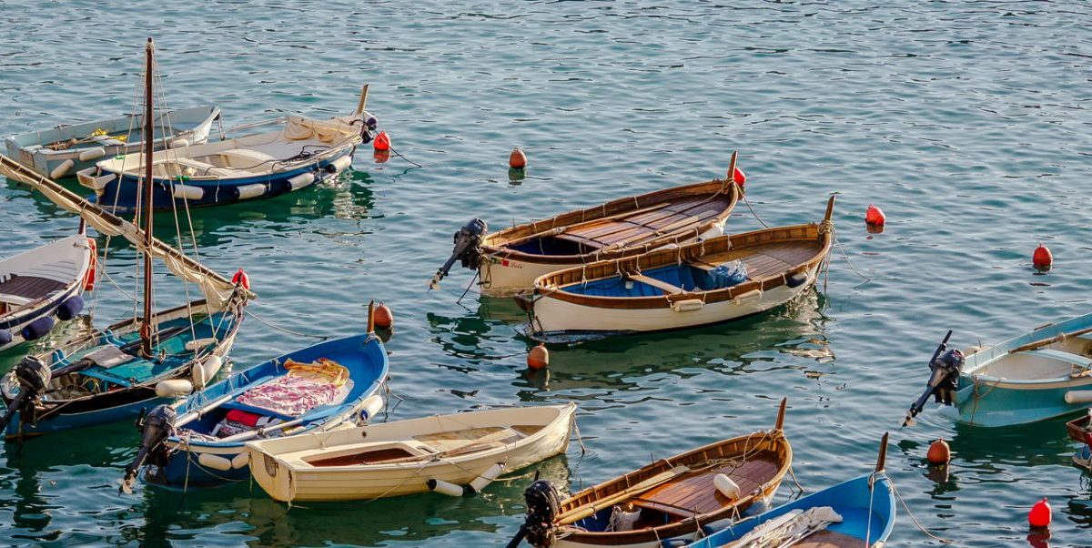 Un grupo de coloridos barcos de pesca flota en el puerto de Vernazza, con aguas azules y tranquilas, boyas rojas y un nadador cerca del borde inferior, debajo del muelle.