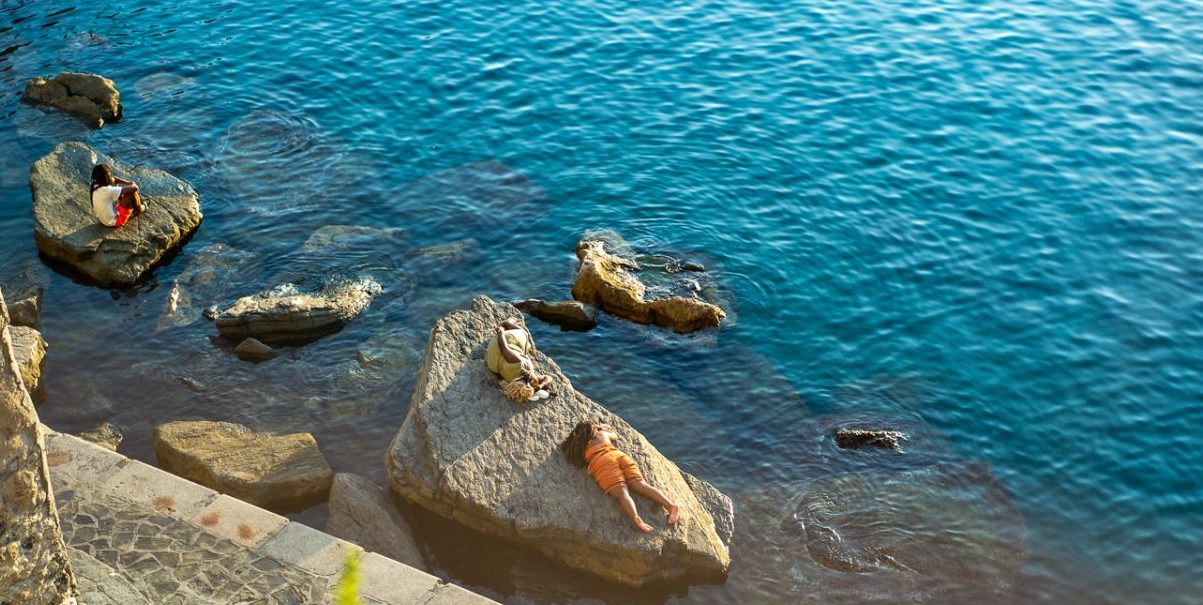 Los bañistas se relajan en las rocas junto al mar debajo del paseo marítimo de piedra en Vernazza, con aguas cristalinas de color azul, barandillas y acantilados iluminados por el sol que enmarcan la cala.